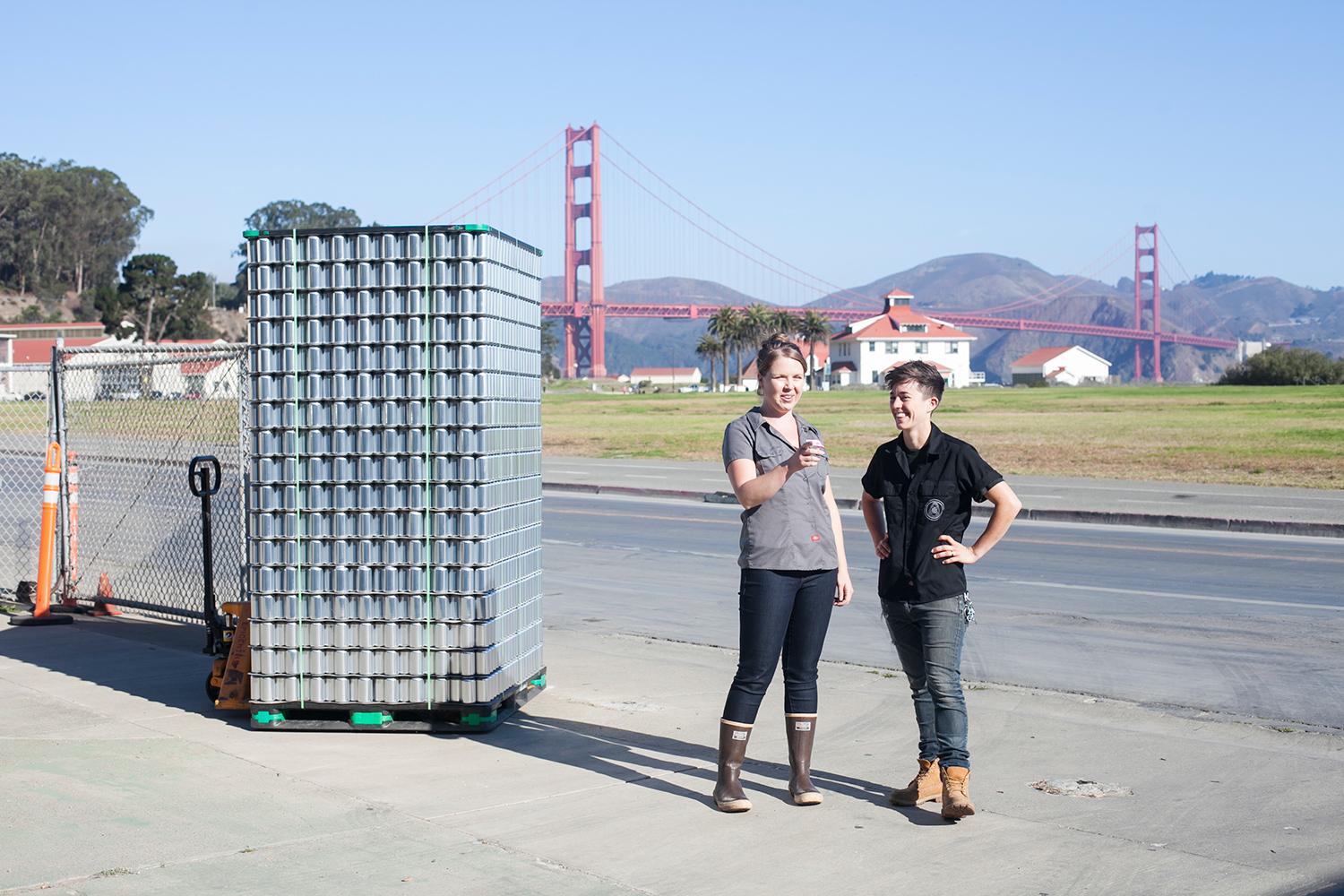 Owners, Jenn and Lindsey, standing in front of the Golden Gate Bridge next to a pallet of empty aluminum cans